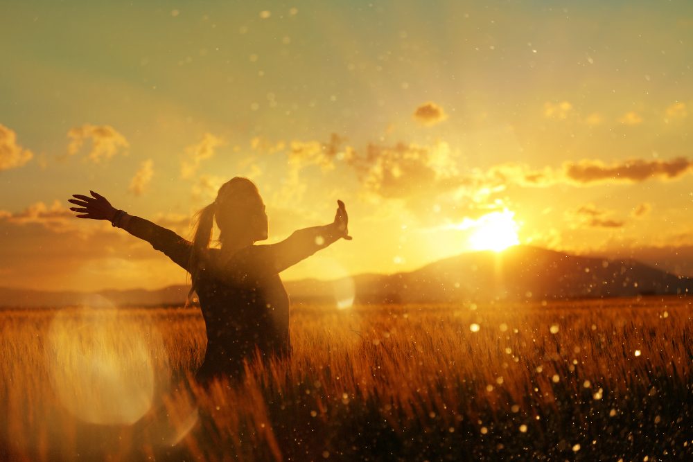 A woman standing in a wheat field at sunrise with her arms open wide